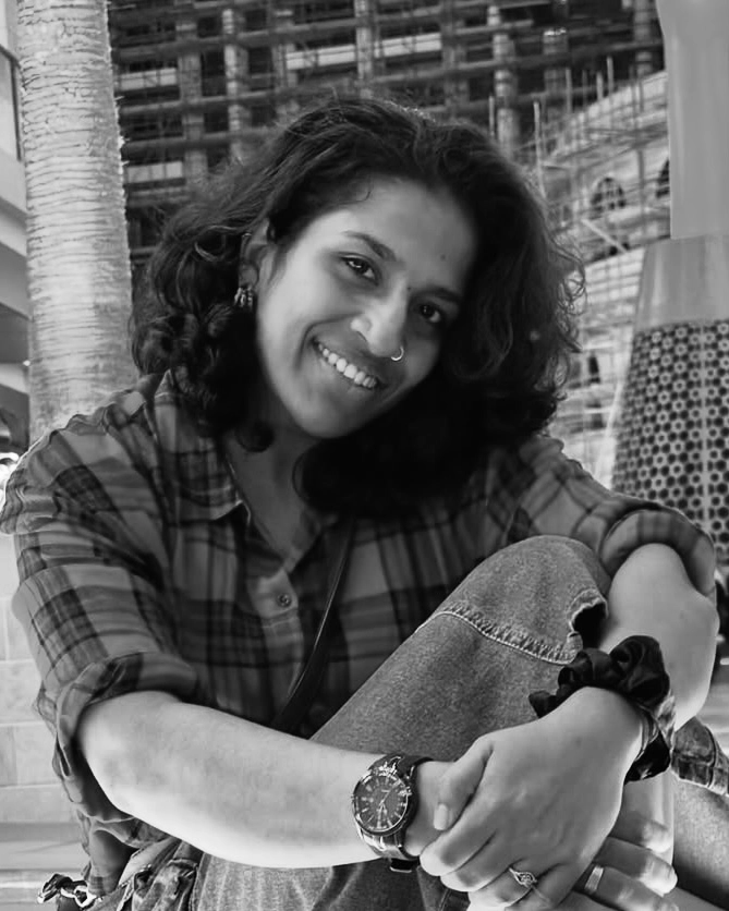 Freelance digital marketer in Qatar A black and white portrait of a person with long ,dark ,curly hair and a warm smile, wearing a plaid shirt and jeans. They are looking directly at the camera.